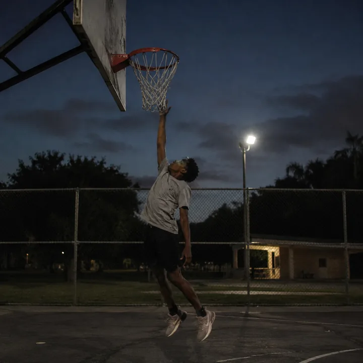 A teenage basketball player jumping to touch the backboard on an outdoor court at dusk